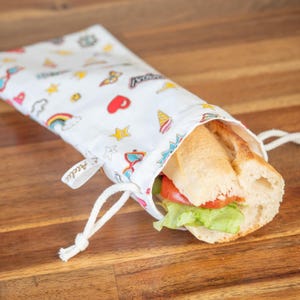 May include: A white fabric sandwich bag with a drawstring closure, patterned with colorful cartoon graphics. The bag holds a baguette sandwich with lettuce and tomato. The bag is on a wooden surface.