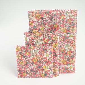 May include: Three reusable food wraps in varying sizes. Each wrap features a vibrant floral pattern with pink, yellow, and white flowers on a red background. The wraps are designed to replace plastic wrap.