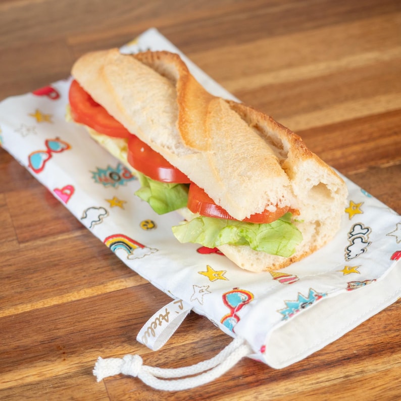 May include: A baguette sandwich with tomato and lettuce on a white, patterned cloth bag. The bag has a drawstring closure and is decorated with colorful cartoon graphics, including rainbows, stars, and sunglasses.