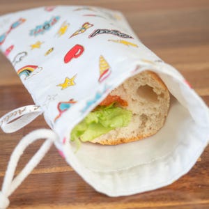 May include: A white fabric sandwich bag with a drawstring closure, filled with a sandwich. The bag is patterned with colorful cartoon graphics. The sandwich contains lettuce and tomato. The bag is open, revealing the sandwich.