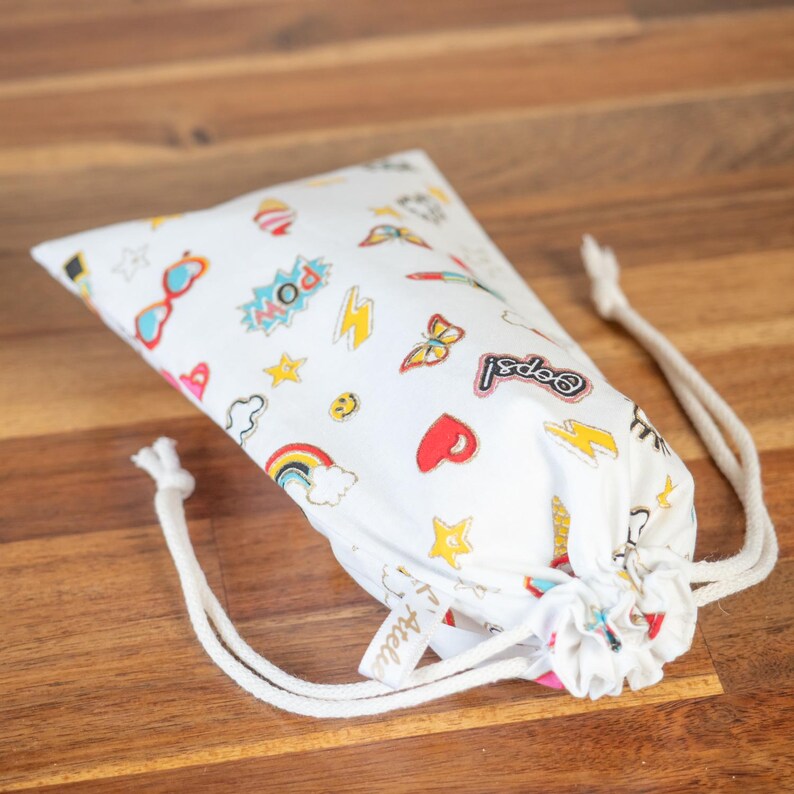 May include: White drawstring bag with a colorful pop art design. The bag features illustrations of sunglasses, lightning bolts, rainbows, stars, hearts, and the word "POW!" in a comic book style. The bag is tied with a white drawstring.