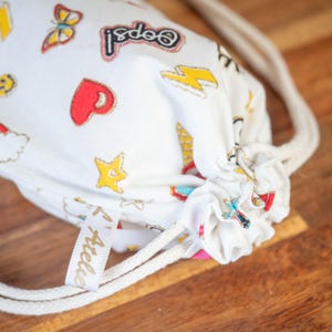 May include: White drawstring bag with colorful cartoon graphics, including a butterfly, heart, star, and lightning bolt. The bag has a cream-colored drawstring and a small tag that says "Atelier".