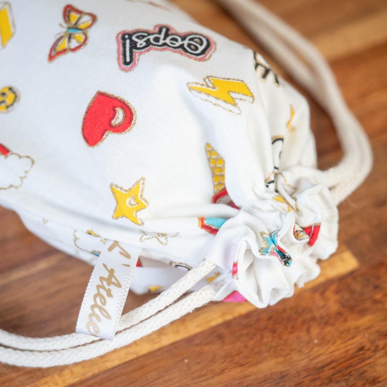 May include: White drawstring bag with colorful cartoon graphics, including a butterfly, heart, star, and lightning bolt. The bag has a cream-colored drawstring and a small tag that says "Atelier".
