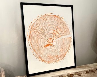 May include: A framed print of a tree ring cross-section in orange on a white background. The print is in a black frame and is sitting on a table with a tiled surface.