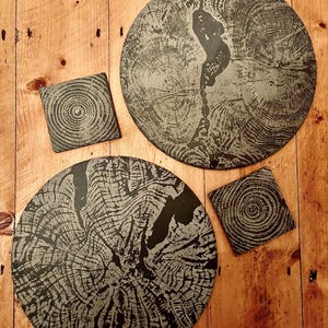 May include: A set of four slate coasters and placemats with a tree ring design. The set includes two large round placemats and two square coasters. The coasters and placemats are gray and black and rest on a wooden surface.