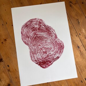 Peut inclure: Impression sur papier blanc représentant un motif de cernes d'arbre bordeaux. Le motif met en valeur les motifs complexes des anneaux de croissance de l'arbre, créant une esthétique naturelle. L'œuvre est posée sur une surface en bois.