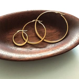 Peut inclure: Trois boucles d'oreilles créoles dorées de différentes tailles, présentées sur une surface en bois.