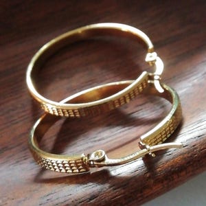 Puede incluir: Un par de pendientes de aro dorados con un diseño texturizado. Los pendientes están hechos de metal y tienen un cierre de bisagra.