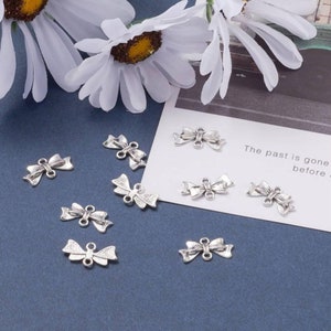 May include: A collection of ten silver-toned bow charms with a textured finish. The charms have two loops on the back for attaching to jewelry or other crafts.