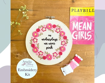 May include: An embroidery kit featuring a floral wreath design with the text "on Wednesdays we wear pink." The wreath is made of pink and red flowers. A "Mean Girls" Playbill and spools of pink thread are also visible.