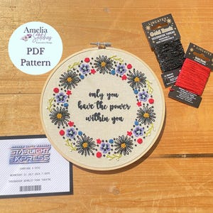 Könnte beinhalten: Ein bestickter Stickrahmen mit einem Blumenkranz und dem Zitat "only you have the power within you". Der Rahmen liegt auf einer Holzoberfläche mit zwei Spulen Stickgarn.