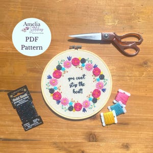 Puede incluir: Un aro de bordado colorido con un diseño floral y el texto "You can't stop the heat!" El aro está sobre una superficie de madera con hilo de bordar y tijeras.