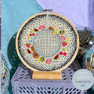 May include: An embroidery kit with a disco ball design. The circular hoop has a cream fabric with a black dotted border and embroidered fruits, lips, and stars. The kit includes the text "Amelia Stitches Embroidery Kit."