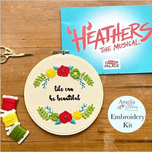 Puede incluir: Kit de bordado para un proyecto temático de "Heathers". El kit incluye un aro con un diseño que dice "la vida puede ser hermosa" con flores y hojas, y tres madejas de hilo de bordar en rojo, amarillo y verde.