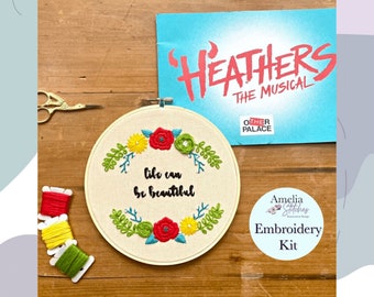 May include: Embroidery kit for a "Heathers" themed project. The kit includes a hoop with a design that says "life can be beautiful" with flowers and leaves, and three skeins of embroidery floss in red, yellow, and green.
