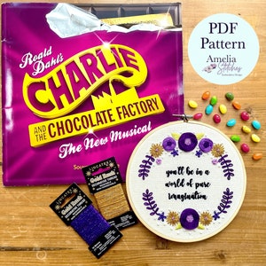 May include: A purple and gold embroidery hoop with the quote "you'll be in a world of pure imagination" surrounded by flowers and leaves. The hoop is on a wooden surface with a book and candy.