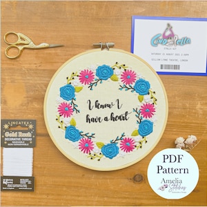 Op de afbeelding: Een witte borduurring met een bloemenkrans en de tekst "I know I have a heart". De ring ligt op een houten oppervlak met een spoel wit garen en een kaartje voor een voorstelling van Assepoester.
