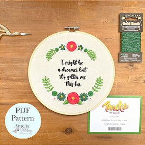 May include: A white embroidery hoop with a floral design and the text "I might be a dreamer but it's gotten me this far". The hoop is on a wooden surface with a green spool of thread and a ticket to the musical "Amelie" on the table.