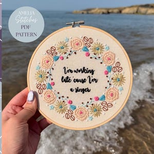May include: Embroidered hoop art with a floral wreath design and the text "I'm working late cause I'm a singer." The embroidery uses pink, blue, and gold thread. The text "Amelia Stitches PDF Pattern" is in the upper left corner.
