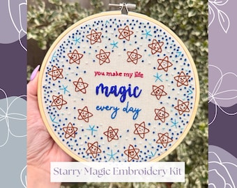 May include: An embroidery hoop with a cream-colored fabric featuring a design of smiling stars and the words "you make my life magic every day." The text and stars are in shades of blue and brown. The kit is labeled "Starry Magic Embroidery Kit."