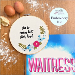 May include: An embroidery kit with the text "she is messy but she's kind" in a wooden hoop, surrounded by floral embroidery. The kit includes a "Waitress" musical playbill, eggs, and flour.