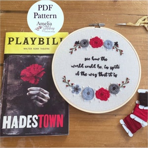 Op de afbeelding: Een witte borduurring met een citaat uit de musical Hadestown: "see how the world could be, in spite of the way that it is". Het citaat is geborduurd met zwart garen, omringd door rode en grijze bloemen. De ring ligt op een houten oppervlak met een programmaboekje voor de musical Hadestown en rode borduurgaren.