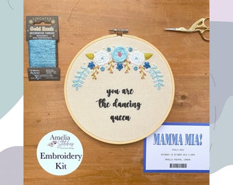 May include: A blue and white embroidery kit with a hoop featuring the text "you are the dancing queen" and a floral design. The kit includes a spool of blue thread and a ticket for the musical Mamma Mia!.