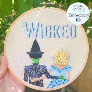 Puede incluir: Arte de bordado en aro con los personajes de Elphaba y Glinda del musical Wicked. El aro es blanco con la palabra "WICKED" bordada en hilo plateado y verde. Los personajes están bordados en detalle, con Elphaba con un vestido verde y Glinda con un vestido azul.