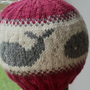 May include: A pink, white, and gray knitted hat with a whale design. The hat is made of wool and has a soft, cozy feel.