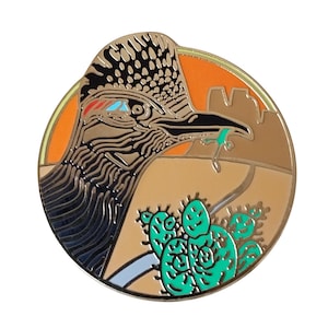 May include: A circular enamel pin featuring a roadrunner with a lizard in its beak. The design includes a desert landscape with a bright orange sky, tan ground, and a vibrant green prickly pear cactus in the foreground, all outlined in gold metal.