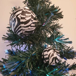 May include: Two black and white zebra print Christmas ornaments hanging on a green artificial Christmas tree with blue and green fiber optic lights.