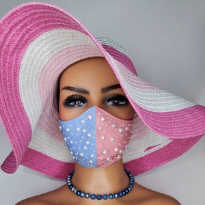 May include: A blue and pink face mask with pearl embellishments. The mask is worn by a mannequin wearing a pink and white striped sun hat and a blue necklace.
