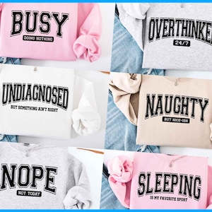 May include: Five different sweatshirts with humorous text printed on the front. The sweatshirts are in various colors, including pink, white, beige, gray, and light pink. The text on the sweatshirts includes "Busy Doing Nothing", "Undiagnosed But Something Ain't Right", "Overthinker 24/7", "Naughty But Nice-ish", "Nope Not Today", and "Sleeping Is My Favorite Sport".
