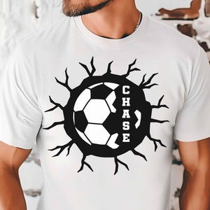 May include: White t-shirt with a black and white soccer ball design. The soccer ball is encircled by a black graphic with the name "CHASE" in white lettering. The design has a cracked, broken effect.
