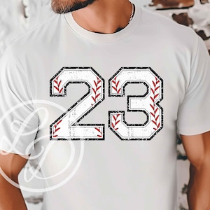 May include: White t-shirt with a distressed black and white number 23. The number is outlined in black and has a baseball stitch design in red.