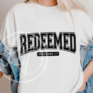 May include: White sweatshirt with the word "REDEEMED" in bold, black, block letters. Below the word is the text "EPHESIANS 1:7" in a smaller, black font. The sweatshirt is worn with a denim jacket.