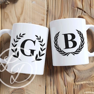 May include: Two white ceramic mugs with black handles. The mugs feature a black wreath design with a letter in the centre. The mug on the left has the letter G and the mug on the right has the letter B.