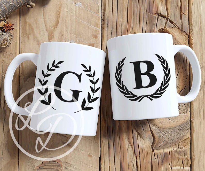 May include: Two white ceramic mugs with black handles. The mugs feature a black wreath design with a letter in the centre. The mug on the left has the letter G and the mug on the right has the letter B.