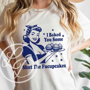 May include: A white t-shirt with a blue graphic of a woman holding a tray of cupcakes. The text on the shirt reads "I Baked You Some Shut The Fucupcakes".