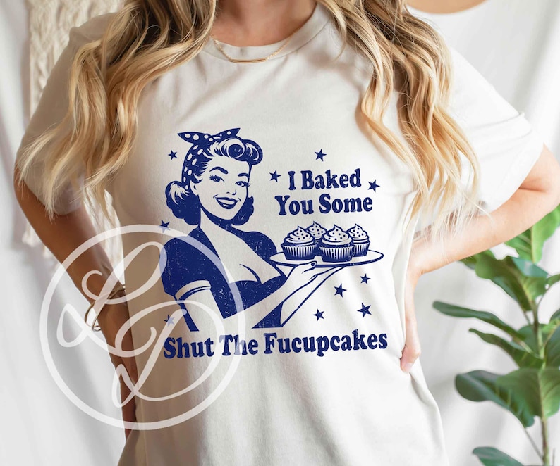 May include: A white t-shirt with a blue graphic of a woman holding a tray of cupcakes. The text on the shirt reads "I Baked You Some Shut The Fucupcakes".