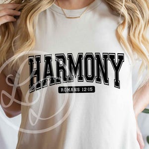 May include: Cream-colored t-shirt with the word "HARMONY" in bold, black, collegiate-style lettering. Below the word is the text "ROMANS 12:15" in a smaller, black font. The shirt is a casual, everyday wear item.