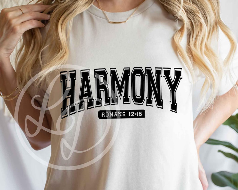 May include: Cream-colored t-shirt with the word "HARMONY" in bold, black, collegiate-style lettering. Below the word is the text "ROMANS 12:15" in a smaller, black font. The shirt is a casual, everyday wear item.