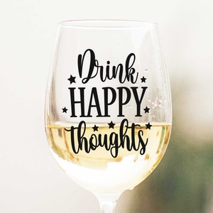 May include: A clear glass wine glass with a black vinyl decal that reads "Drink Happy Thoughts" with stars around the text.