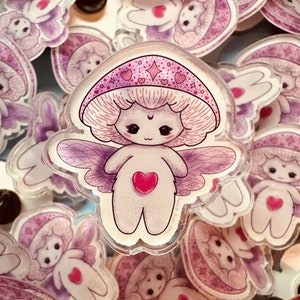 Puede incluir: Una colección de pegatinas transparentes de hadas de hongos acrílicos. Las pegatinas presentan un diseño caprichoso con un sombrero de hongo, alas y un corazón en el pecho. La paleta de colores incluye tonos de rosa, morado y blanco, con contornos negros. Las pegatinas están dispersas sobre una superficie.