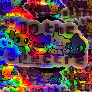 Op de afbeelding: Holografische sticker met vier stripfiguren en de tekst "ON THE SPECTRE" en "Angelic Angel Gifts". De sticker heeft een regenboogglans en is omgeven door soortgelijke stickers.
