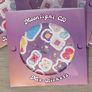 Puede incluir: Una hoja de pegatinas con forma de CD con varios diseños coloridos, incluyendo corazones, estrellas y una cara de gato, es visible a través de un paquete morado y rosa. El paquete dice "Moonlight CD" y "Mix Stickers" en una fuente decorativa.