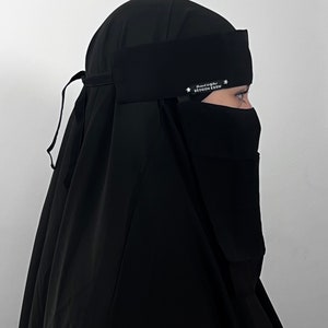 Könnte beinhalten: Schwarzer Niqab mit schwarzem Gesichtsschleier und einem schwarzen Stirnband mit dem Text "SEDQON ESSM" in weißer arabischer Schrift und einem weißen Stern auf jeder Seite.