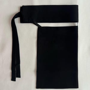 May include: A black head covering featuring a rectangular panel and a wide band. The band has two long, thin ties extending from the side. The head covering is made of a smooth, opaque fabric.
