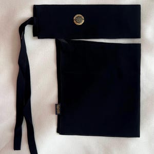 Peut inclure: Un bandeau de tête en tissu noir avec un emblème doré. L'enveloppe a une partie supérieure rectangulaire avec un emblème doré circulaire et de longues attaches. Un morceau de tissu rectangulaire est attaché en dessous. L'étiquette sur le tissu indique "myoto".