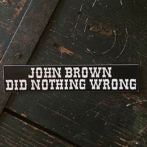 May include: A black rectangular sticker with the text "JOHN BROWN DID NOTHING WRONG" in white, bold, capital letters. The sticker is placed on a dark, weathered wooden surface with metal accents.